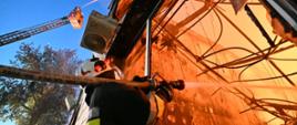 Strażak wyposażony w wąż strażacki z dyszą, gasi ogień wewnątrz płonącej hali strumieniem wody, przez rozbite okno. Sam stoi na zewnątrz. W tle widać wysięgnik wozu strażackiego z platformą, z której również leje się strumień wody, na wyższe piętro hali.
