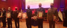Przedstawiciele służb stojący na scenie filharmonii trzymający w dłoniach pamiątkowe ramy z podziękowaniami. W środku zdjęcia stoi w mundurze wyjściowym st. bryg. Jacek Kaczmarek - Kujawsko-Pomorski Komendant Wojewódzki PSP