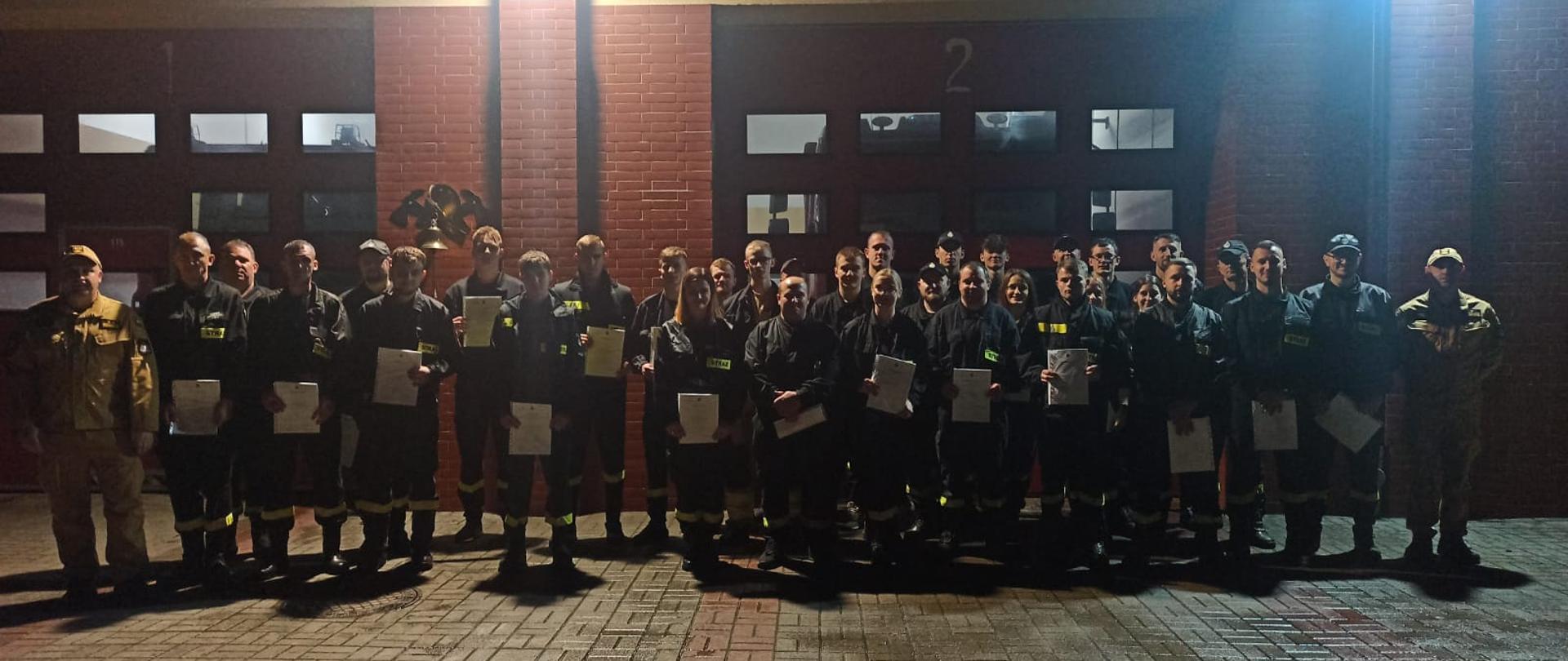 Na placu Komendy Powiatowej Państwowej Straży Pożarnej w Rypinie stoją przed garażami strażacy, którzy zdali kurs podstawowy. Razem z nimi są: dowódca jednostki ratowniczo-gaśniczej brygadier Dariusz Łęgosz wraz ze swoim zastępcą młodszym brygadierem Cezarym Nowińskim.