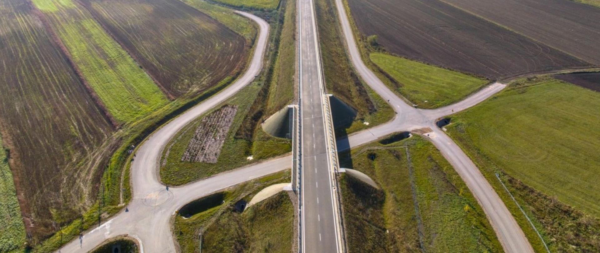 Widok z lotu ptaka na wiadukt drogowy nad lokalną drogą, otoczony polami uprawnymi i drogami dojazdowymi. 