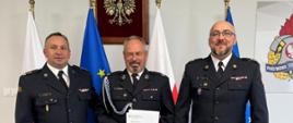 Komendant Powiatowy PSP w Pucku bryg. Wojciech Wanat wraz z Zastępcą kpt. Krzysztofem Mingą wręczają promesę prezesowi OSP Strzelno Joachimowi Marzejon. Wszyscy są ubrani w mundury galowe. Za mężczyznami znajdują się na flagi Rzeczpospolitej Polskiej oraz Unii Europejskiej oraz Godło Polski. Po prawej widnieje logo Państwowej Straży Pożarnej.