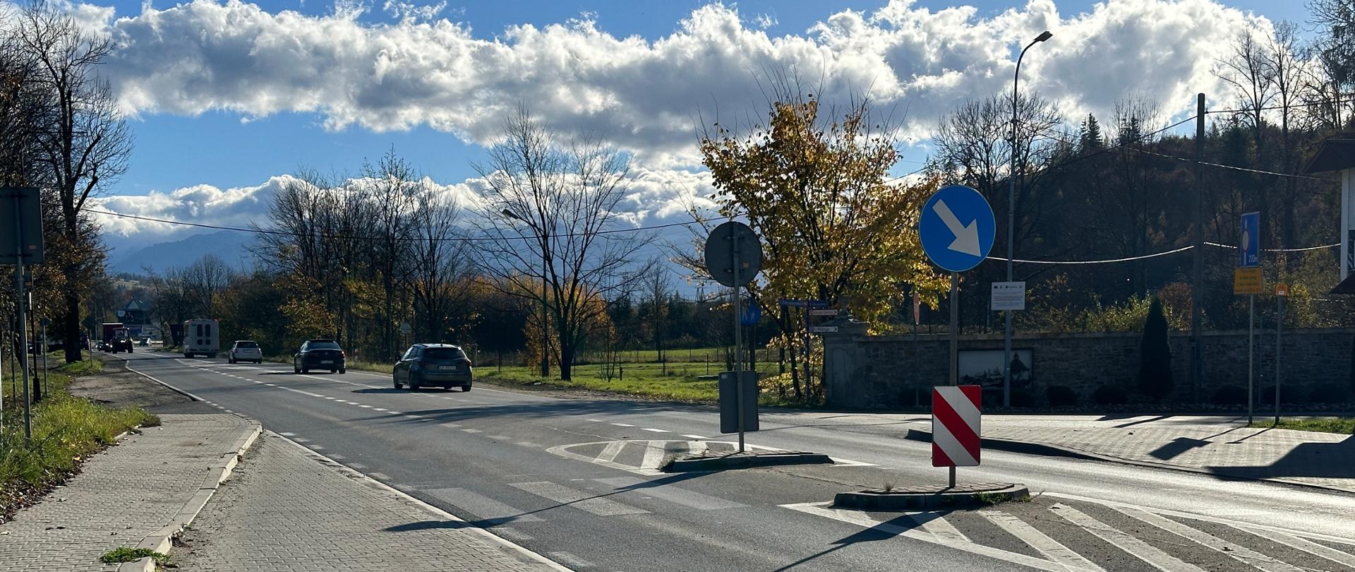 DK47 Zakopianka w Szaflarach, droga jednojezdniowa, na pierwszym planie chodnik i zatoka autobusowa, na jezdni przejście i wyspa azylu, na drodze samochody, w tle zadrzewienia