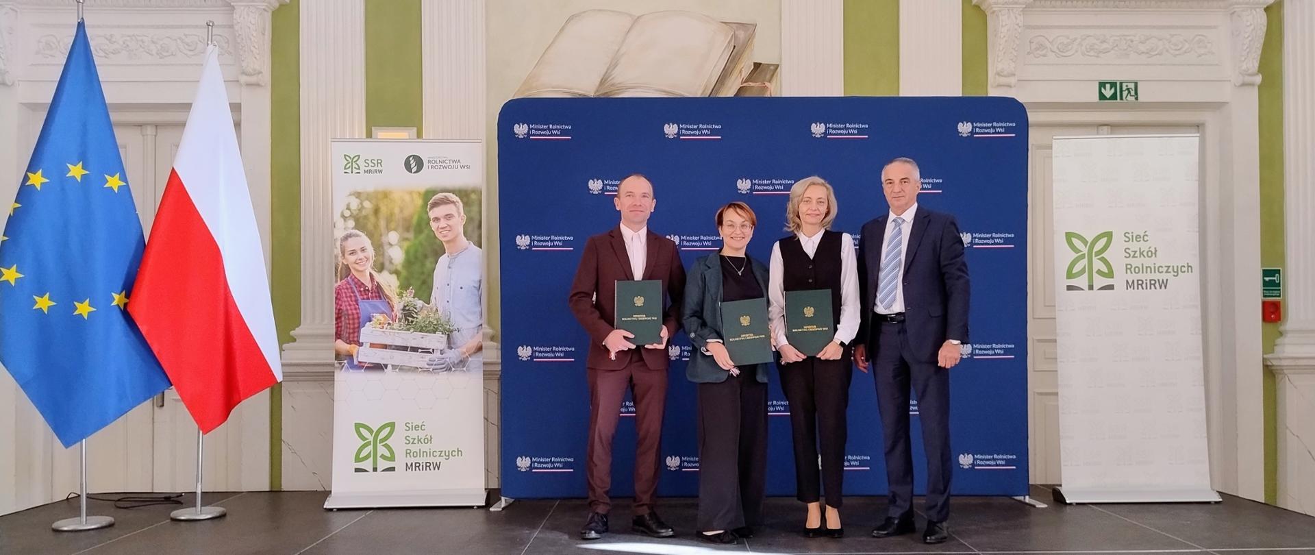 Cztery osoby stoją na tle banerów MRiRW i sieci szkół rolniczych, trzy prezentują otrzymane akty nadania stopnia awansu zawodowego
