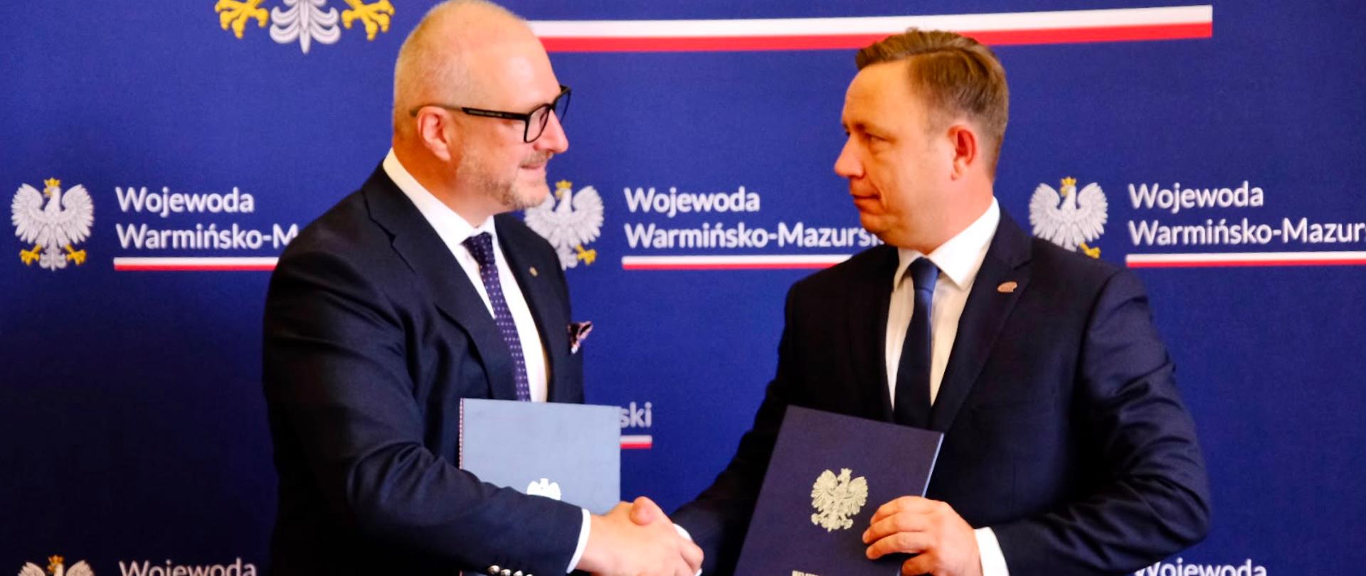 Dwóch mężczyzn w granatowych garniturach stoi na tle ścianki Województwa Warmińsko-Mazurskiego i podaje sobie ręce, patrząc na siebie.