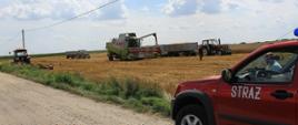 Maszyny rolnicze podczas prac żniwnych. Na pierwszym planie widoczne auto operacyjny straży pożarnej, w tle kombajny wysypujący ziarno do przyczep.