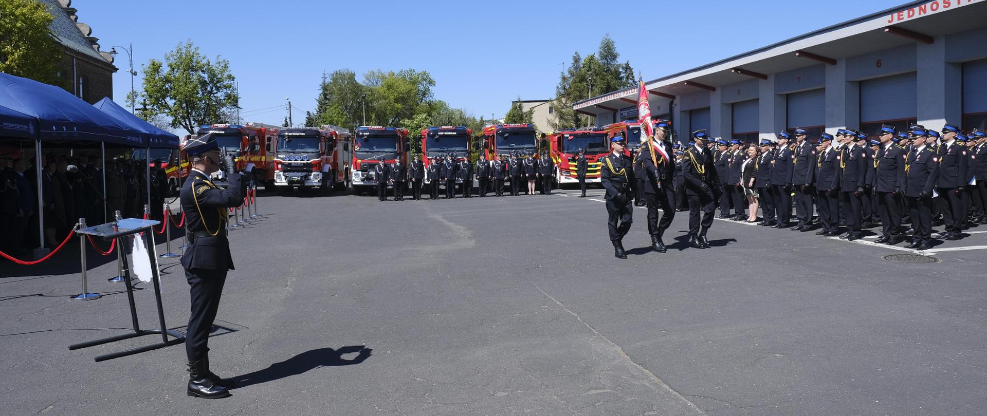 Na zdjęciu pododdziały strażaków w umundurowaniu wyjściowym ustawione na placu przed jednostką Ratowniczo-Gaśniczą nr II podczas uroczystej zbiórki. przed frontem pododdziałów maszeruje poczet sztandarowy.