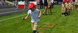 XI Powiatowe Zawody Sportowo-Pożarnicze OSP i MDP. Stadion w Miejskiej Górce. Na specjalnym torze przeszkód prezentują się członkowie DDP. Jedna z zawodniczek biegnie po ułożonej na murawie drabince. Z tyłu na swoją kolejnośc startu oczekują pozostałe koleżanki i koledzy. W tle namioty.
