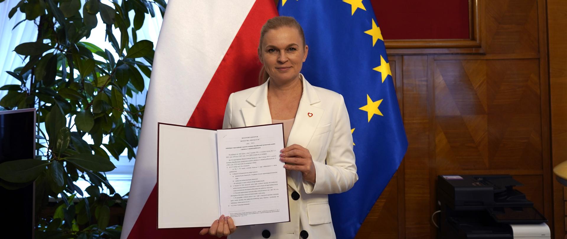 Minister Barbara Nowacka stoi na tle flagi Polski i Unii Europejskiej. W ręku trzyma otwartą teczkę, w której znajduje się podpisane rozporządzenie o pracach domowych. W tle na ścianie w drewnianej ramie na czerwonym tle wisi orzeł, symbol Polski. 