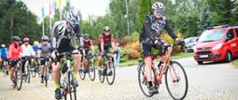 Grupa kolarzy-strażaków rusza na trasę pielgrzymki, jadąc po kostce brukowej przed kościołem. Na pierwszym planie rowerzysta w sportowym stroju z elementami niebieskiego i pomarańczowego oraz białym kasku. Za nim jadą kolejni uczestnicy. W tle widoczny jest budynek kościoła, flagi i czerwony samochód strażacki.