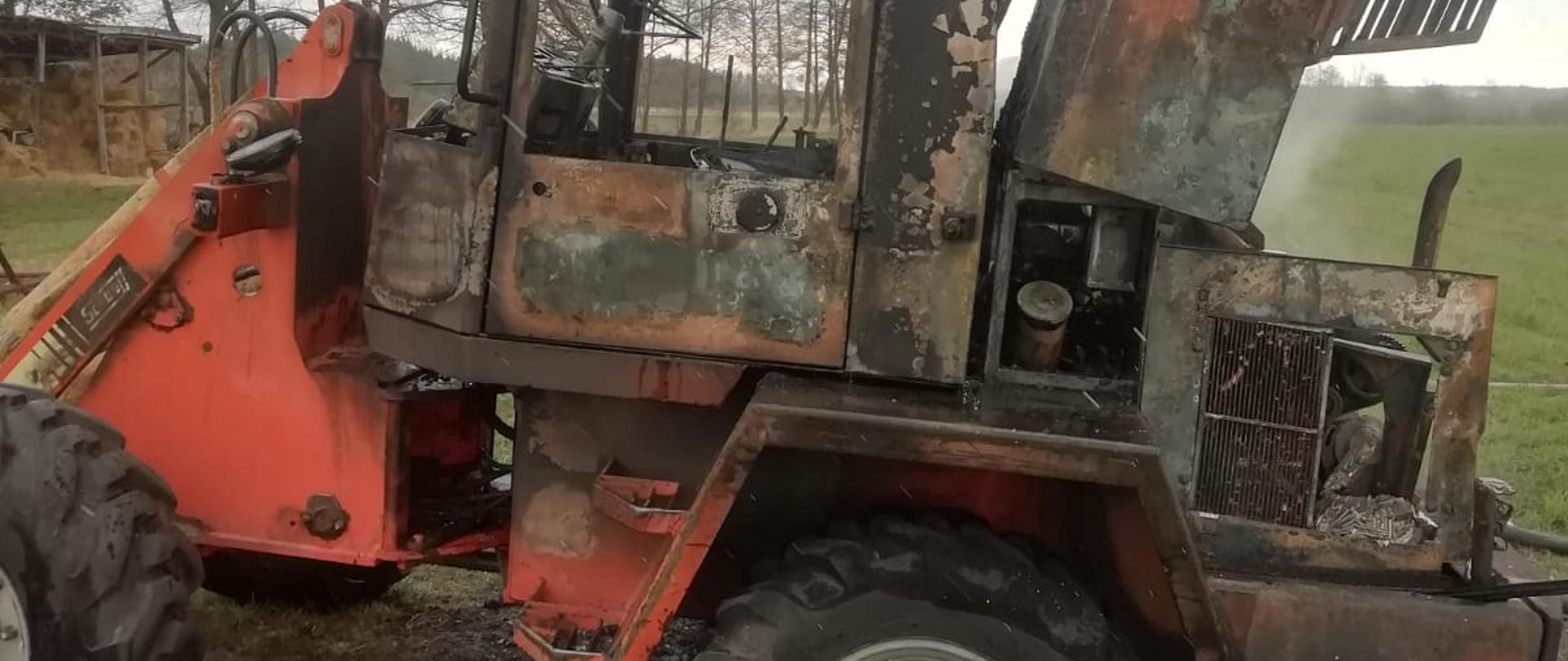 Na zdjęciu ładowarka po pożarze. Spaleniu uległa komora silnika oraz kabina. Zdjęcie wykonane z lewego boku pojazdu, który znajdował się na jezdni. W tle łąka i drzewa.