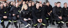 na sali siedzą na krzesłach ratownicy osp ubrani w czarne ubrania koszarowe w tle w drewnianej gablocie sztandar komendy powiatowej psp tablica pamiątkowa oraz tablo z pracownikami komendy