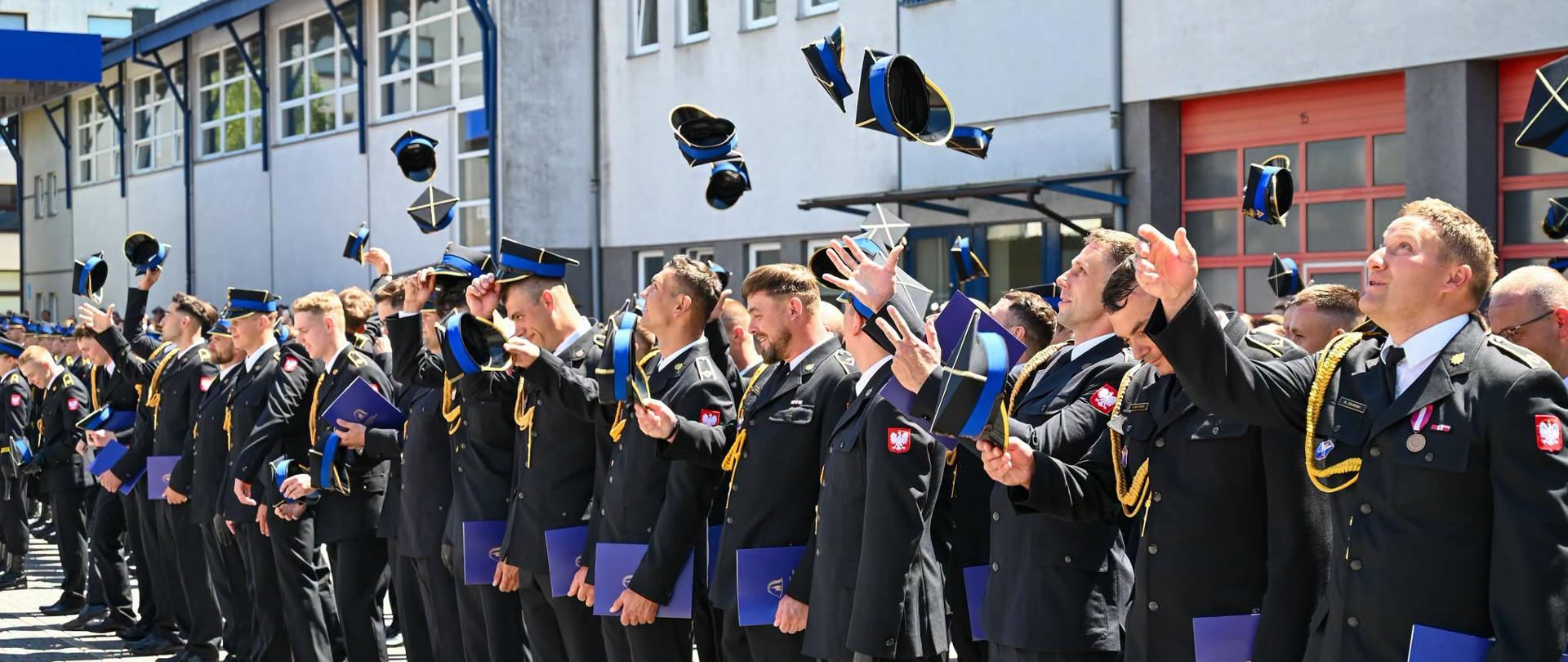 Grupa absolwentów w mundurach Państwowej Straży Pożarnej stoi w szeregu na placu przed budynkiem jednostki. Strażacy podrzucają w górę czapki rogatywki i trzymają w rękach niebieskie teczki z dokumentami. Zdjęcie przedstawia radosny moment zakończenia uroczystości promocyjnej.