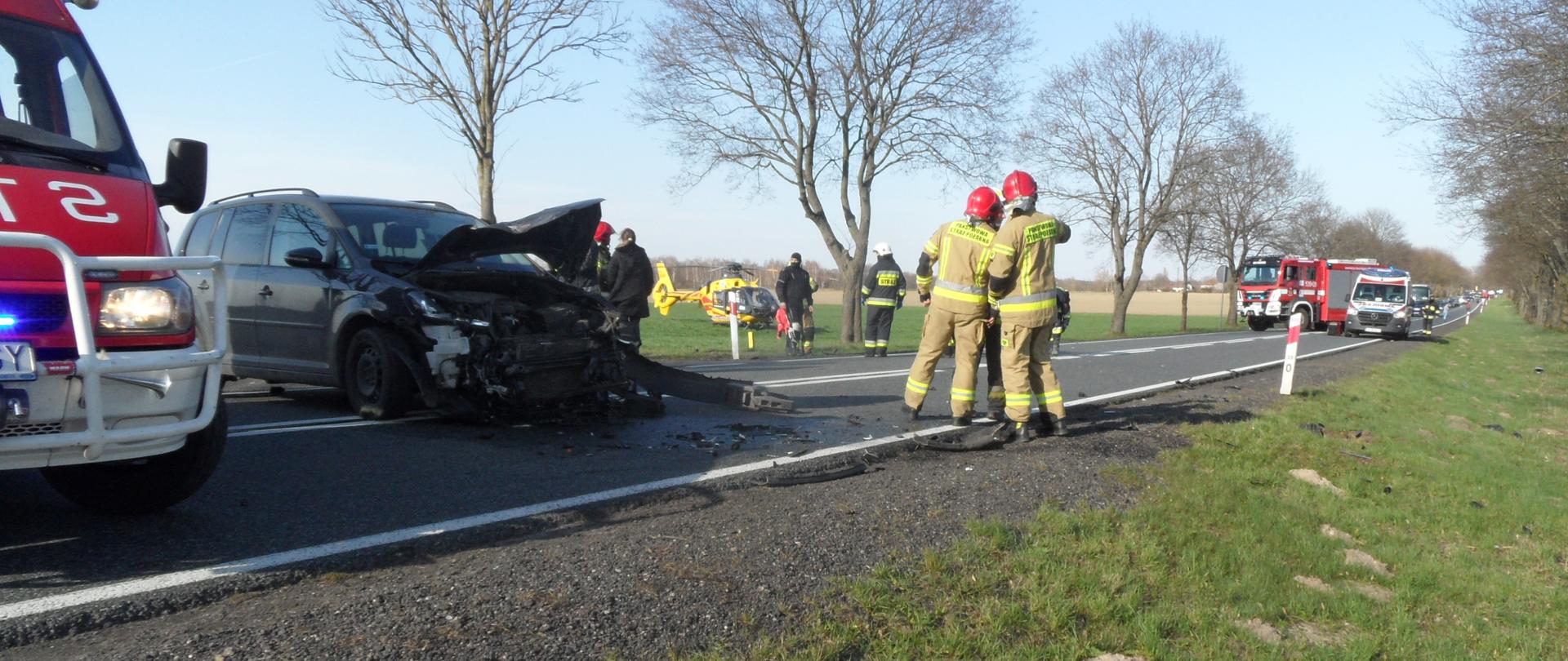 Rozbity samochód na drodze. Dalej samochód straży pożarnej i karetka pogotowia, na polu widać również śmigłowiec LPR