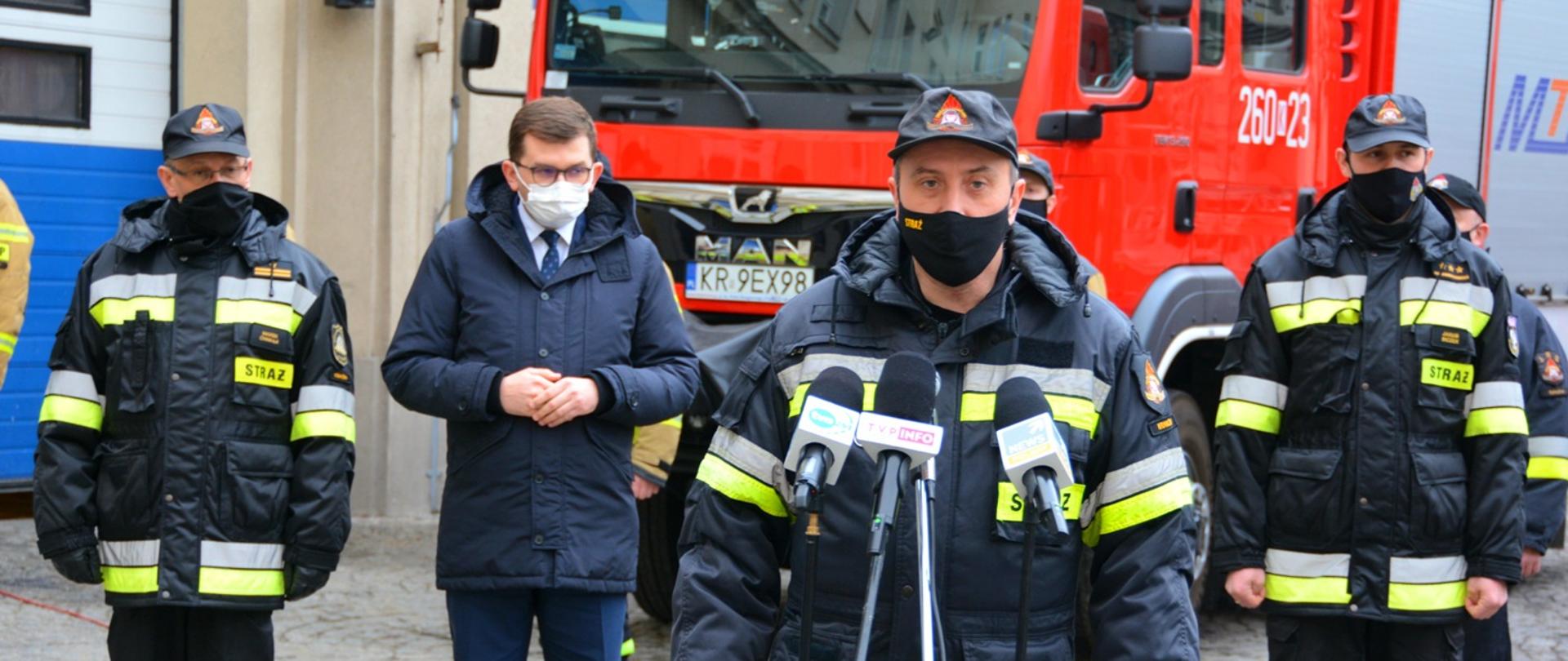 Odprawa z przygotowującymi się do wyjazdu strażakami. Głos zabiera małopolski komendant wojewódzki PSP. W tle stoją strażacy i wojewoda małopolski..