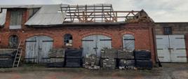 Zdjęcie przedstawia budynek gospodarczy pozbawiony do połowy pokrycia dachowego, widać konstrukcję dachu w postaci łat drewnianych, część dachu zabezpieczona taśmami przed obsunięciem się w dół. Niebo ciemnoszare, deszczowe