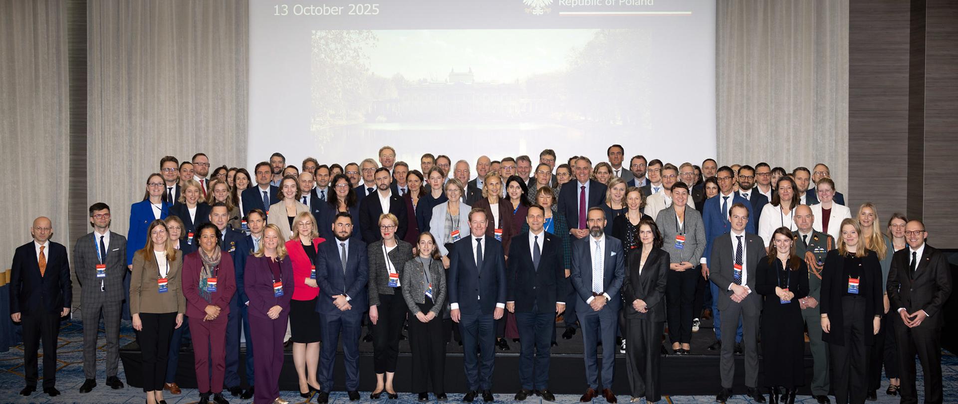 Na zdjęciu jest grupa kilkudziesięciu osób. Stoją w kilku rzędach. Patrzą w kierunku kamery. Są ubrani w ciemne, eleganckie stroje. Za nimi jest duży ekran z napisem XXXIV Utrecht Conference
