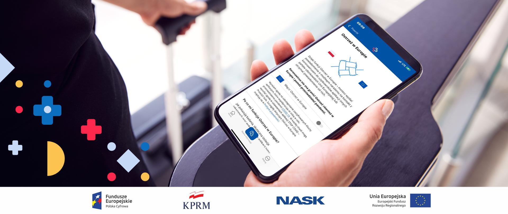 Na pierwszym panie telefon komórkowy. Na jego ekranie wyświetla się aplikacja STOP COVID - ProteGO Safe i jej funkcja "Ostrzeż w Europie". Telefon jest trzymany przez osobę, która wybiera się w podróż. Na drugim planie - widoczna walizka.