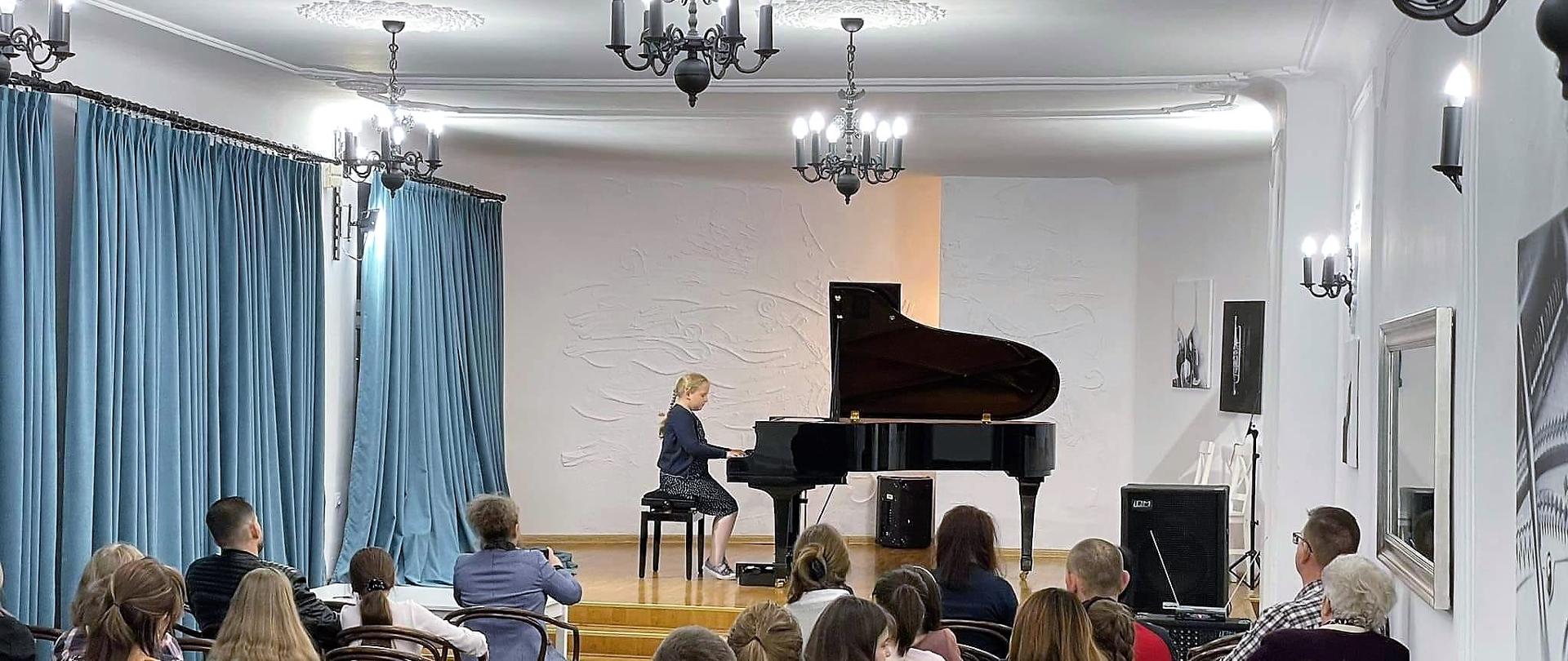 Na zdjęciu znajduje się sala koncertowa, na scenie przy czarnym fortepianie siedzi dziewczynka ubrana w granatowy sweter i czarno-białą spódnicę. Podłoga sceny to jasne drewno, tło stanowi biała ściana, zasłony w kolorze niebieskim, żyrandole w kolorze czarnym. 