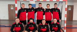 Drużyna strażaków Komendy Powiatowej Państwowej Straży Pożarnej w Białobrzegach pozująca na hali sportowej po zakończonych rozgrywkach futsalu. Zawodnicy uśmiechają się do aparatu, stojąc przed bramką.