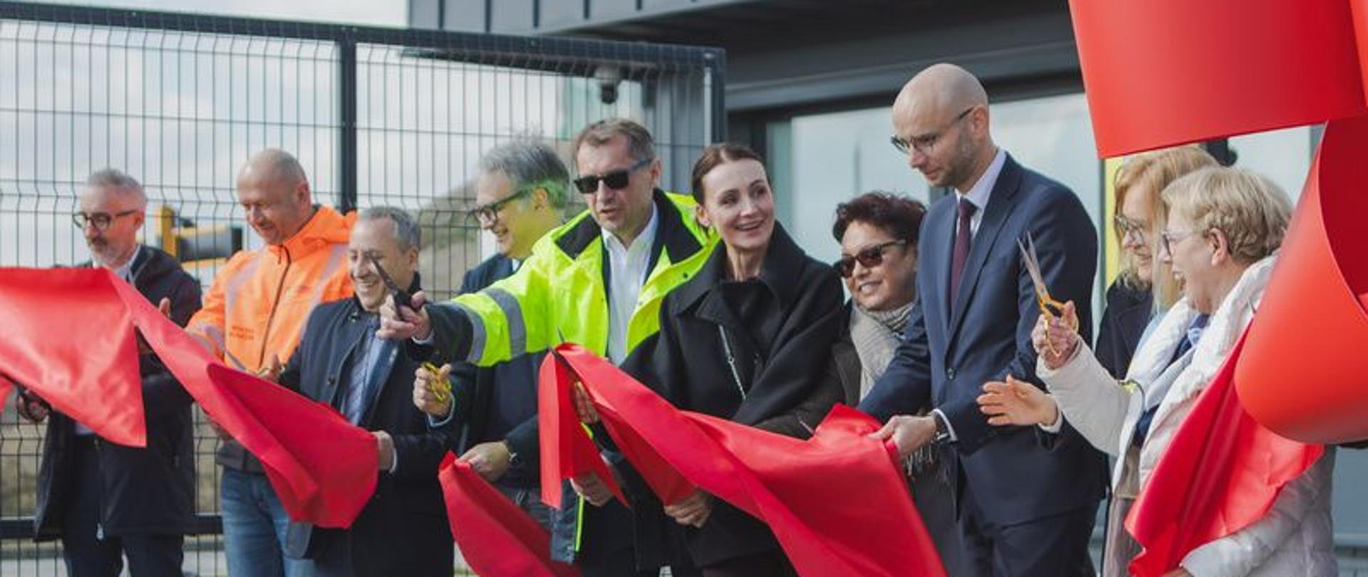 Na zdjęciu widzimy grupę osób uczestniczących w uroczystym przecięciu czerwonej wstęgi — symbolicznym otwarciu obiektu. W tle znajduje się nowoczesny budynek oraz ogrodzenie z metalowej siatki. Część osób trzyma nożyczki i przecina wstęgę, uśmiechając się.