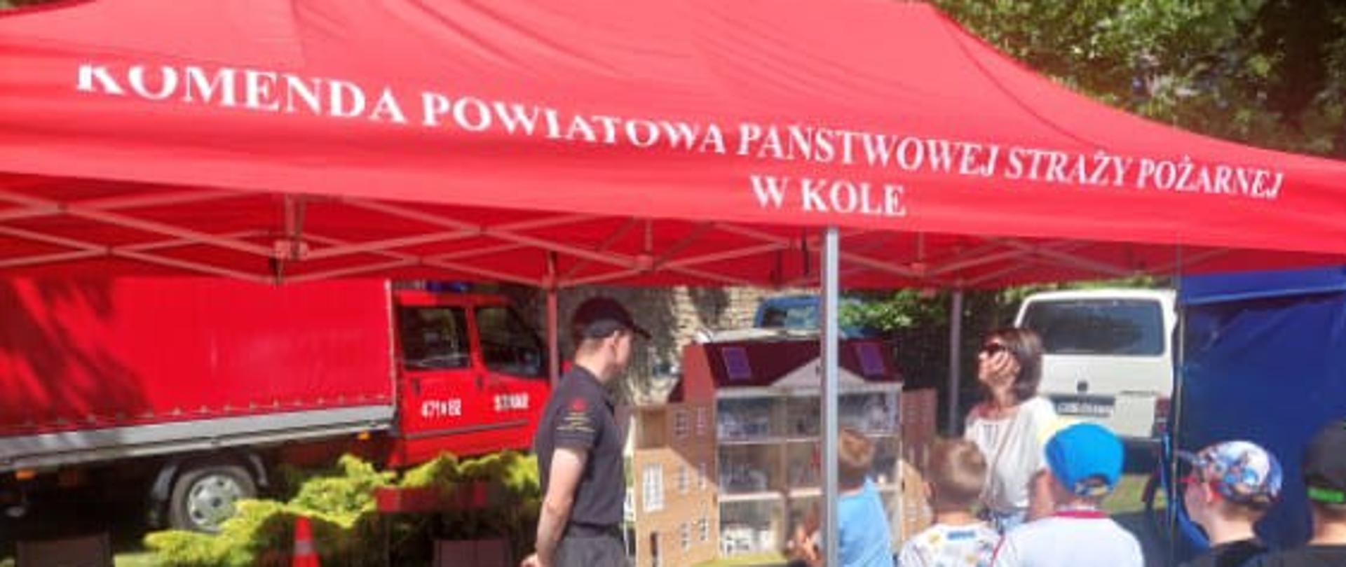 Na zdjęciu widoczny jest namiot z czerwonym zadaszeniem, na którym widnieje napis: „KOMENDA POWIATOWA PAŃSTWOWEJ STRAŻY POŻARNEJ W KOLE”. Pod namiotem znajduje się strażak w mundurze, który rozmawia z grupą dzieci oraz osobą dorosłą. Przed nimi znajduje się makieta domu, prawdopodobnie służąca do edukacji na temat bezpieczeństwa pożarowego. Na trawie, obok namiotu, leżą dwa fantomy do nauki pierwszej pomocy (resuscytacji krążeniowo-oddechowej). W tle widoczne są wozy strażackie oraz inne pojazdy. Całość odbywa się w pogodny dzień, na świeżym powietrzu, prawdopodobnie podczas wydarzenia edukacyjnego lub pikniku rodzinnego.
