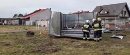 Na zdjęciu strażacy ubrani w ubrania specjalne koloru czarnego oraz hełmy koloru białego i żółtego, zabezpieczają garaż blaszany przewrócony przez wiatr. Garaż znajduje się na trawie, w tle widać budynki, kojec do psa oraz ogrodzenia.