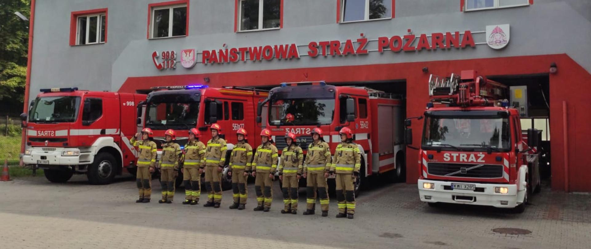 Strażacy w szyku w umundurowaniu bojowym przed samochodami pożarniczymi