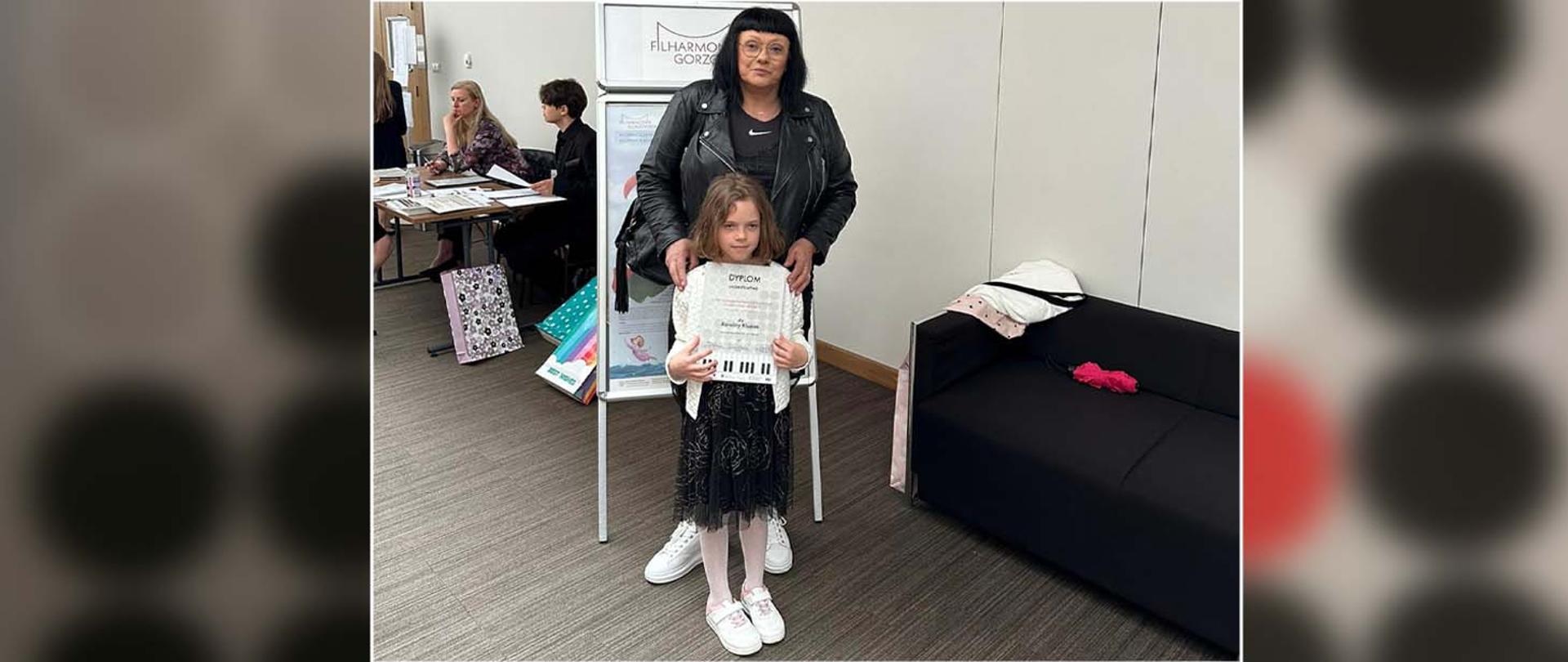 Z przodu nauczycielka z uczennicą, uczennica pokazuje otrzymany dyplom, z tyłu przy stole siedzą inne osoby.