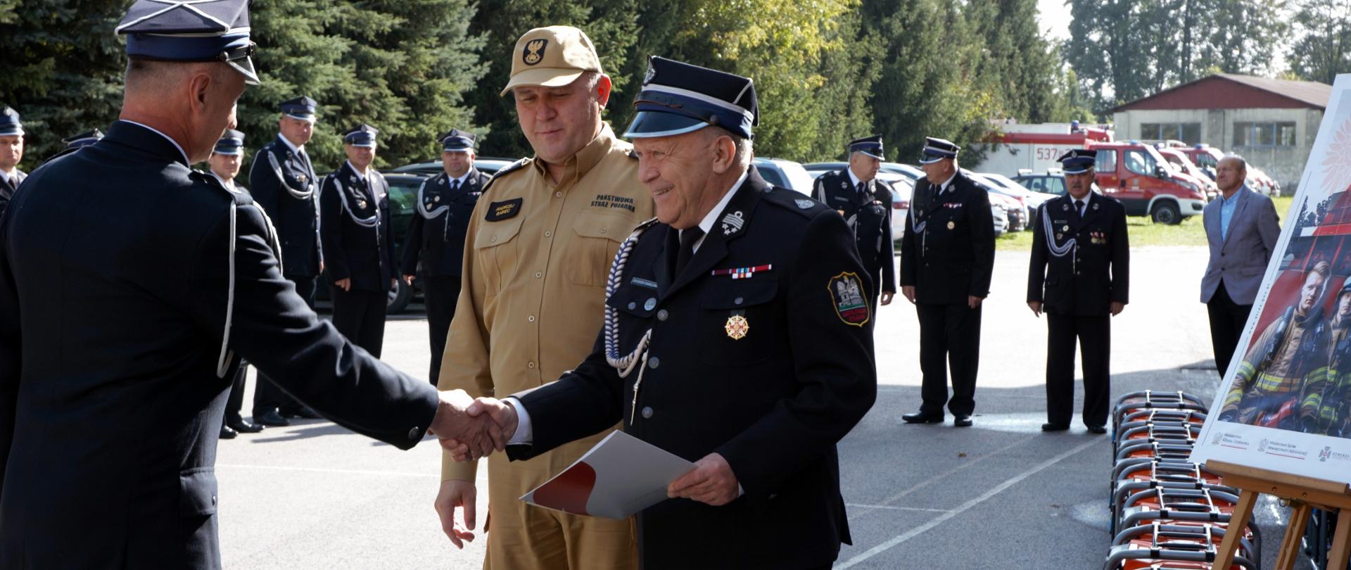 Na asfaltowym placu na pierwszym planie dwóch strażaków (jeden PSP w mundurze w kolorze piaskowym, drugi w mundurze OSP) stoją naprzeciw trzeciego (w mundurze OSP). Jeden strażak OSP przekazuje dokumenty drugiemu strażakowi OSP podając mu przy tym rękę. Za nimi inni strażacy w mundurach OSP ustawieni w szeregu. Po prawej stronie zdjęcia ustawione agregaty prądotwórcze oraz na stojaku plakat. W tle zaparkowane samochody OSP i PSP oraz drzewa.