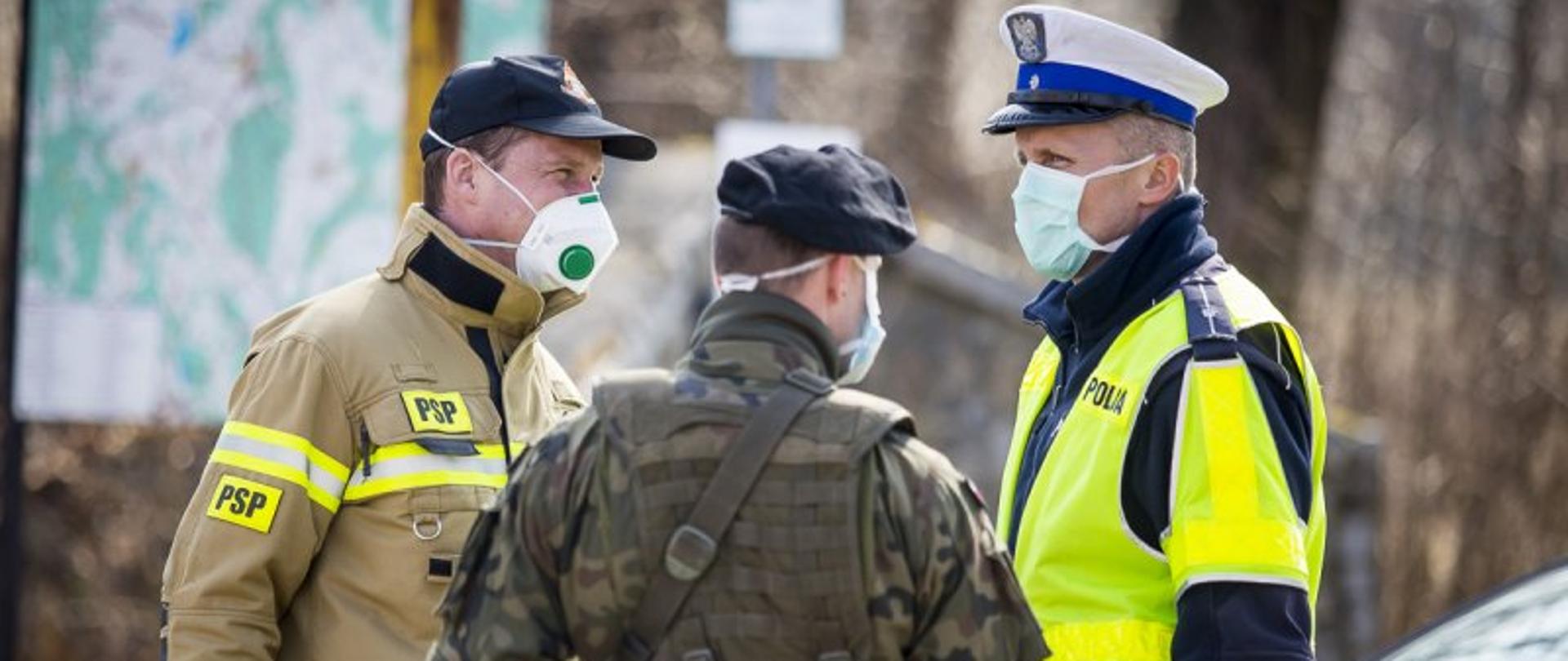 Działania Strażaków Państwowej Straży Pożarnej z Komendy Powiatowej PSP w Zgorzelcu oraz strażaków z OSP KSRG – na zdjęciu przedstawiciele trzech służb, w tym strażak PSP. Rozmawiają w maseczkach.