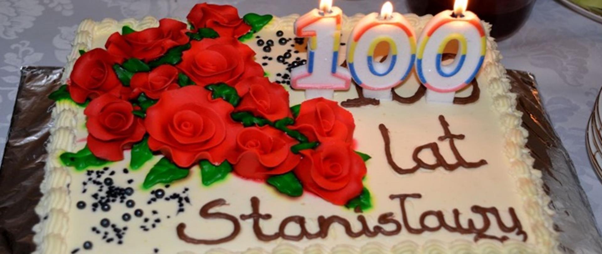 Na zdjęciu znajduje się tort z napisem 100 lat Stanisławy. Tort jest koloru białego z umieszczonymi na nim czerwonymi różami.
