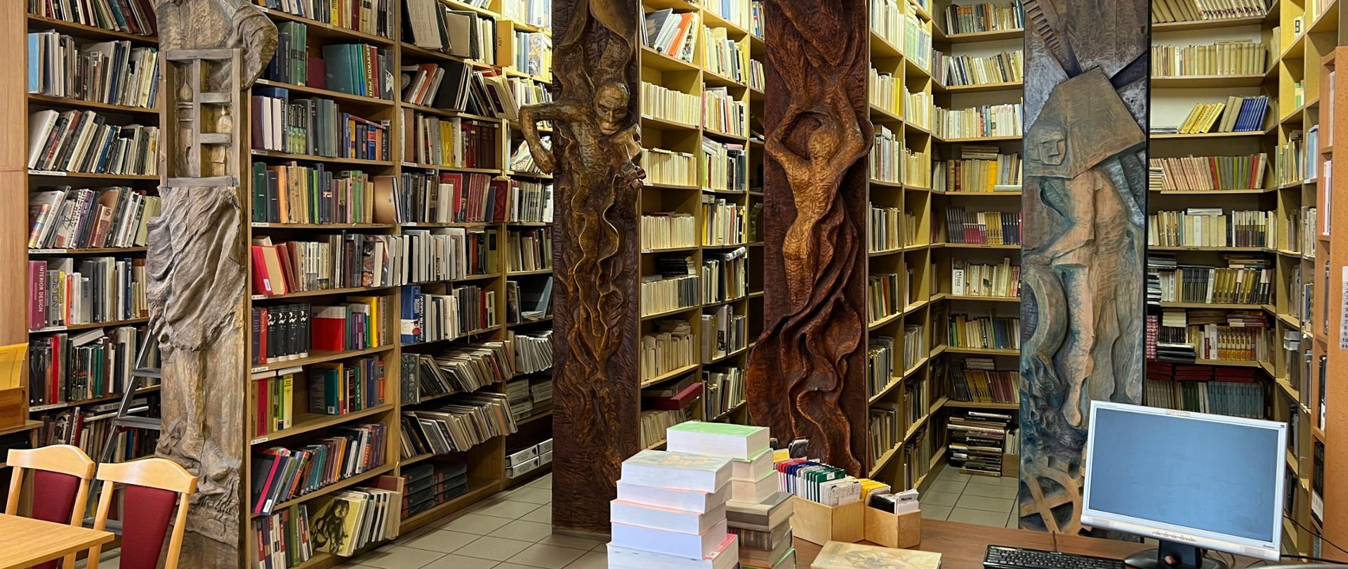 Zdjęcia przedstawia wnętrze biblioteki. Na dole kadru widoczny od lewej: fragment stolika z krzesłami oraz biurko z książkami i komputerem. W głębi regały ze zbiorami książek. Regały na bokach mają ozdobne płaskorzeźby.