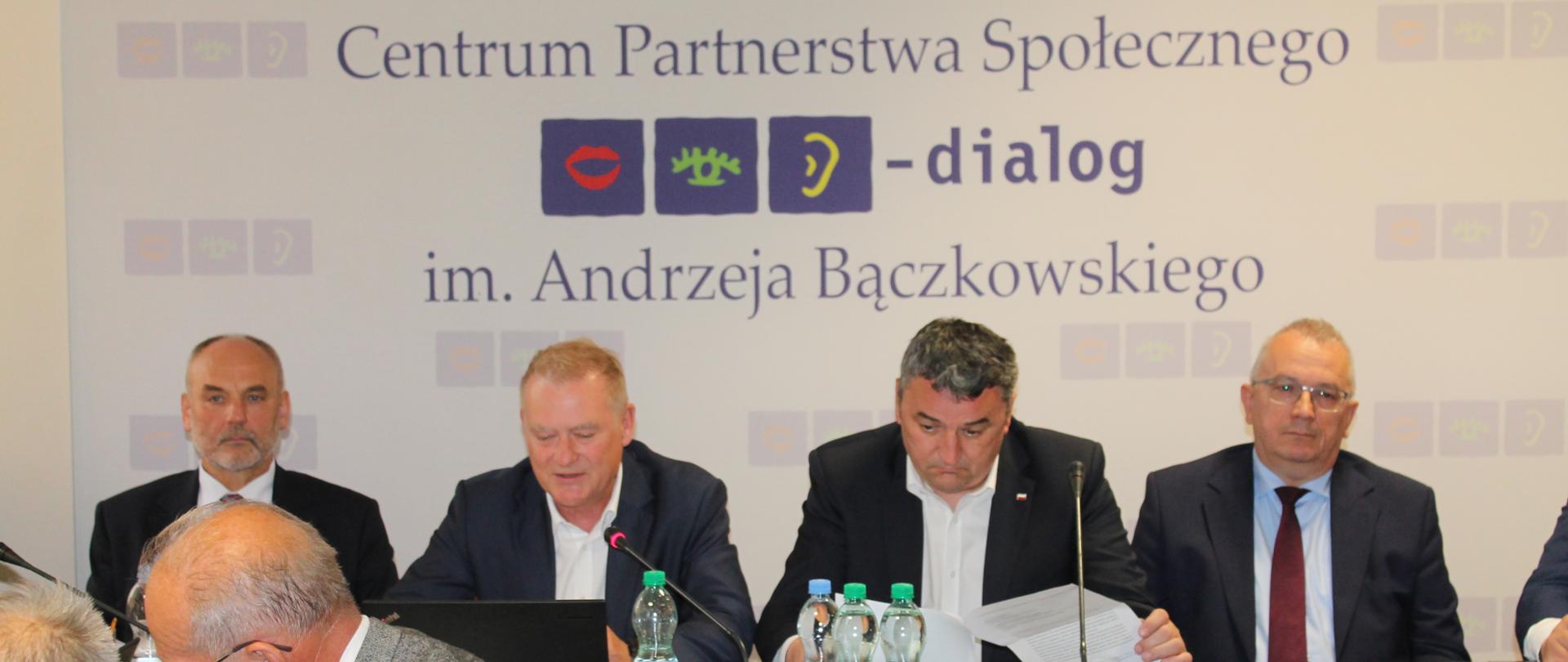 Na zdjęciu widnieje stół prezydialny za którym siedzą: Pan Marek Wesoły, Sekretarz Stanu w Ministerstwie Aktywów Państwowych, Pan Piotr Dziadzio, Podsekretarz Stanu w Ministerstwie Klimatu i Środowiska, Pan Krzysztof Kisielewski, Współprzewodniczący Zespołu Trójstronnego ds. Branży Energetycznej ze strony związków zawodowych oraz Pan Przemysław Kołodziejak, Współprzewodniczący Zespołu Trójstronnego ds. Branży Energetycznej ze strony pracodawców. W tle znajduje się baner z napisem Centrum Partnerstwa Społecznego "Dialog" im. Andrzeja Bączkowskiego.