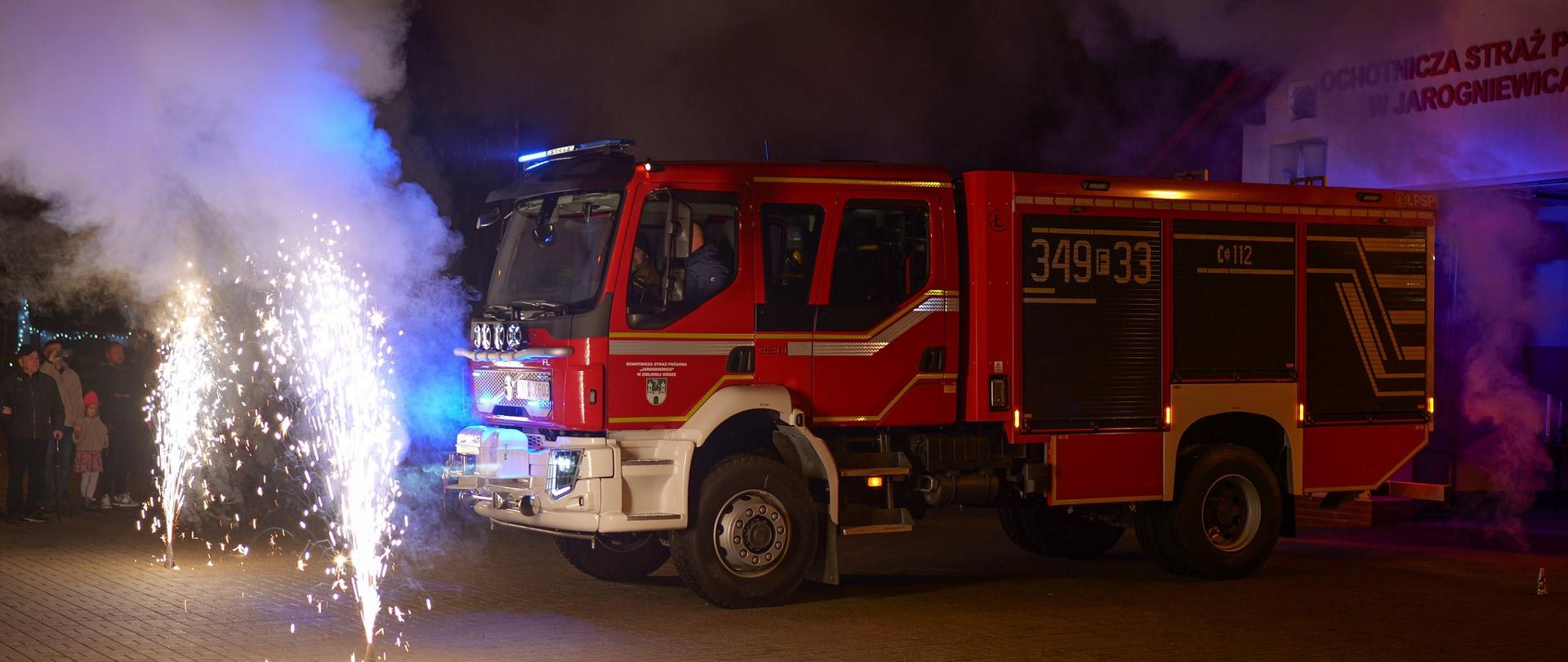 Nowy średni samochód ratowniczo-gaśniczy OSP Jarogniewice (numer boczny 349F33) zaparkowany nocą przed remizą. Z lewej strony odpalone są race świetlne, sugerujące uroczyste powitanie pojazdu.