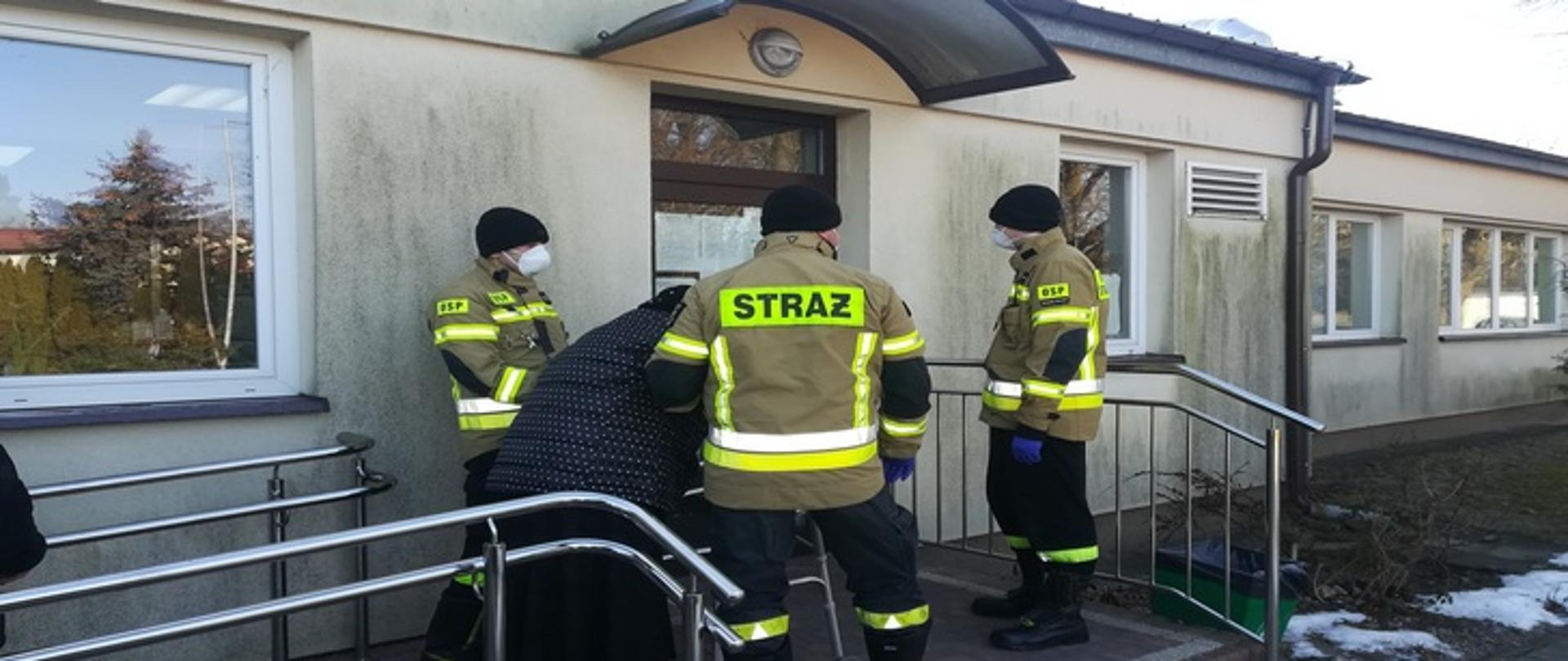 Zdjęcie przedstawia trzech strażaków prowadzących starszą kobietę do wejścia do budynku Ośrodka Zdrowia.
