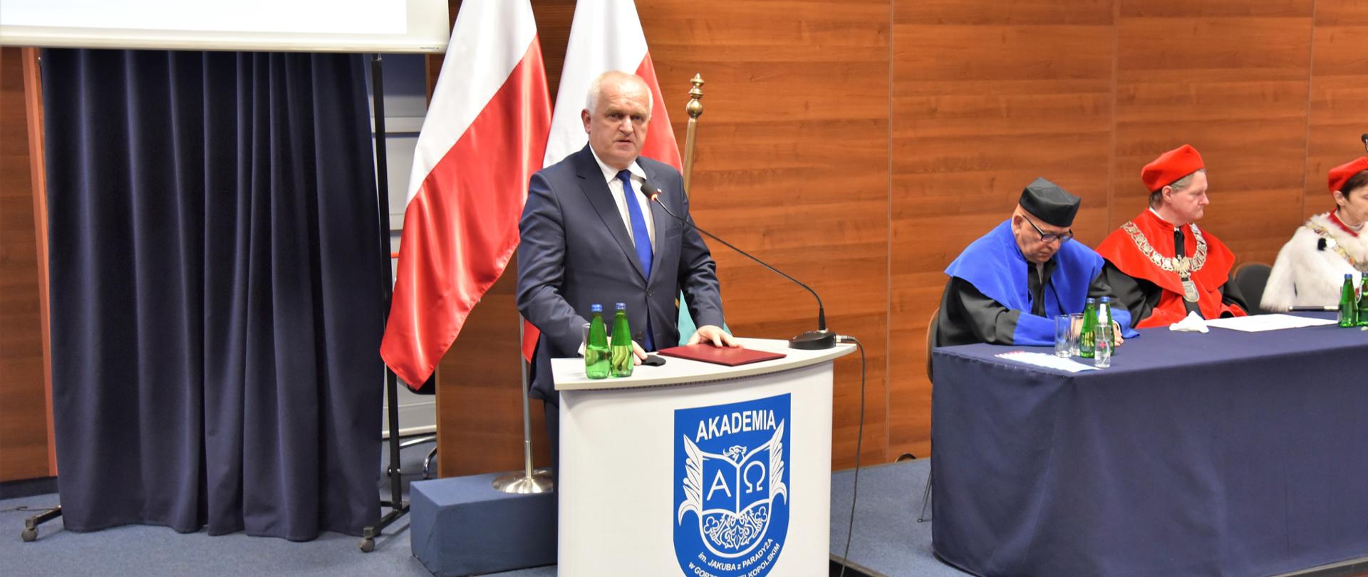 Wojewoda przemawia w czasie uroczystego rozpoczęcia roku, stojąc przy mównicy z logo Akademii. W tle fragment stołu prezydialnego z siedzącymi za nim członkami senatu ubranymi w togi