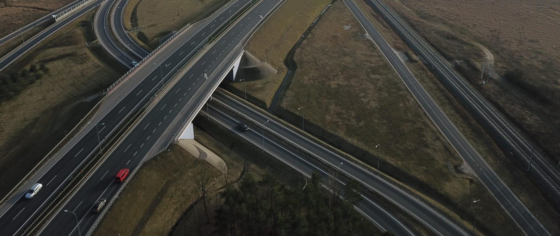 węzeł drogowy w ciągu S11 z lotu ptaka. Droga główna dwujezdniowa przedstawiona w centralnym miejscu przebiega na wiadukcie, pod którym zlokalizowane są drogi dojazdowe do drogi głównej. Po prawej stronie widoczna droga serwisowa i linia kolejowa. Po lewej stronie w oddali widoczne zabudowania. 