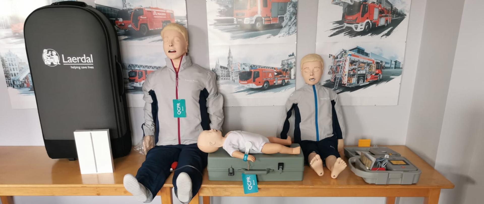 Zdjęcie przedstawia kompletny zestaw do szkolenia z zakresu kwalifikowanej pierwszej pomocy. Na zdjęciu znajdują się: fantom osoby dorosłej, fantom dziecka, fantom niemowlaka oraz pojemniki do przechowywania sprzętu.