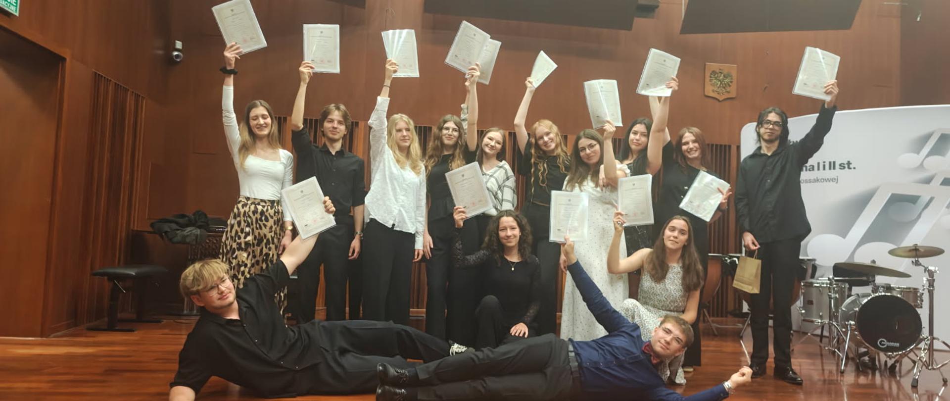 Absolwenci II st. 2026 na scenie w sali koncertowej szkoły. 15 absolwentów z dyplomami w rękach