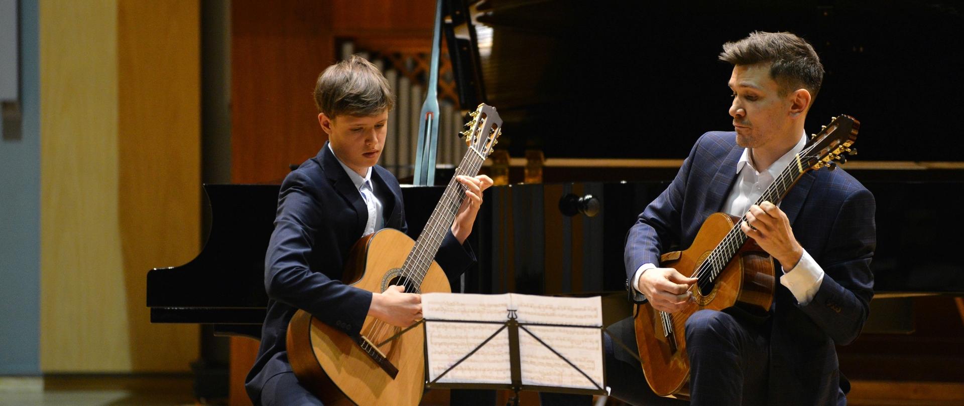 Mistrz i uczeń. Duet gitarowy grający na scenie sali koncertowej. Przed nimi pulpit, na którym ustawione są nuty. Gitarzyści w pozycji siedzącej. Za nimi fortepian, w tle organy.