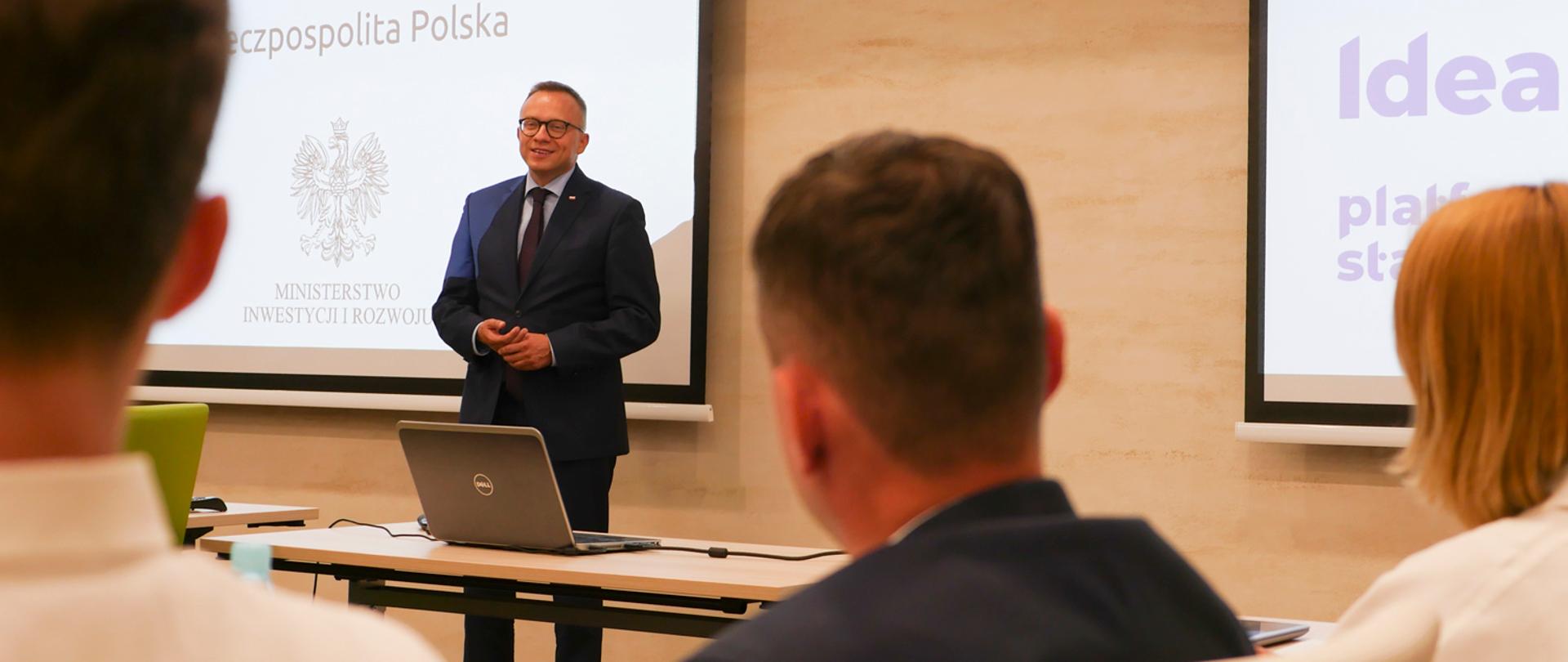 wiceminister Artur Soboń stoi na tle prezentacji, z przodu zdjęcia widoczni patrzący na ministra słuchacze