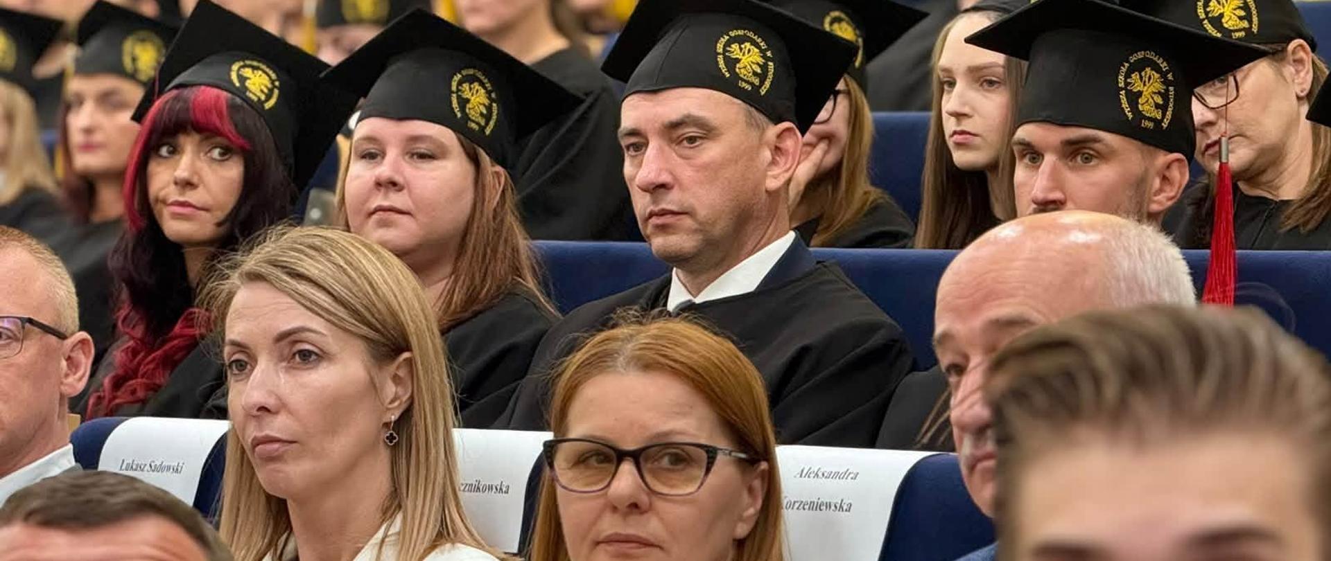 Grupa absolwentów siedzi na widowni. Absolwenci mają na sobie czarne czapki absolwenckie i czarne peleryny.