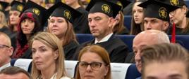 Grupa absolwentów siedzi na widowni. Absolwenci mają na sobie czarne czapki absolwenckie i czarne peleryny.