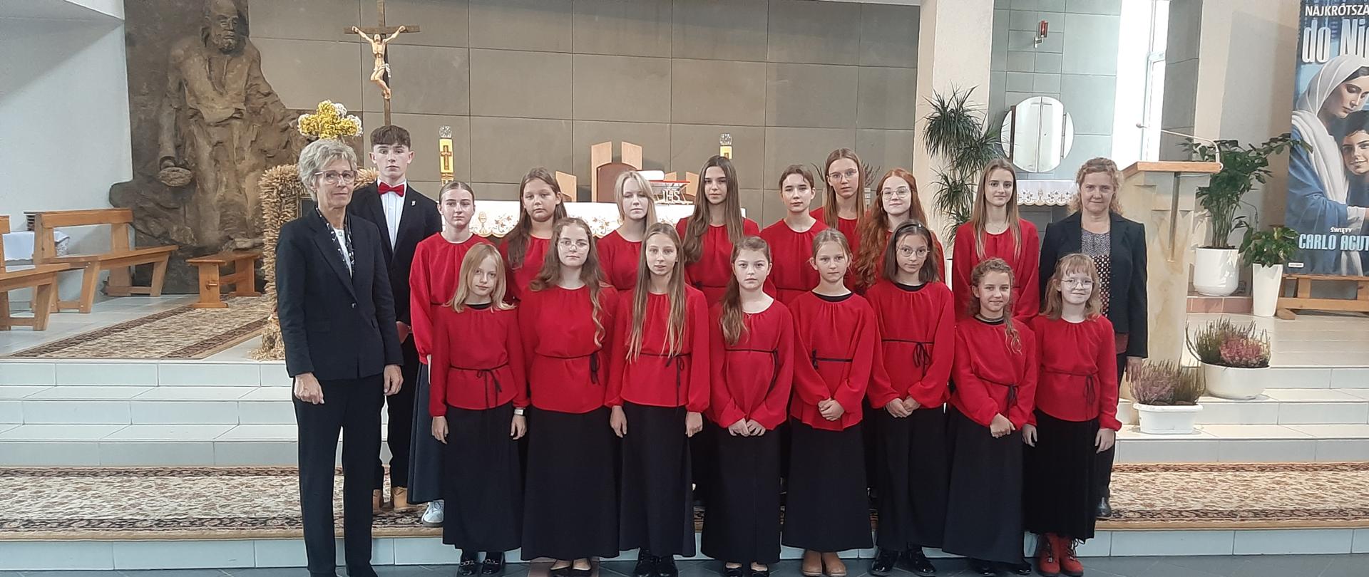 Młodzieżowy zespól wokalny z dyrygentem i akompaniatorem stoi w kościele na tle ołtarza.