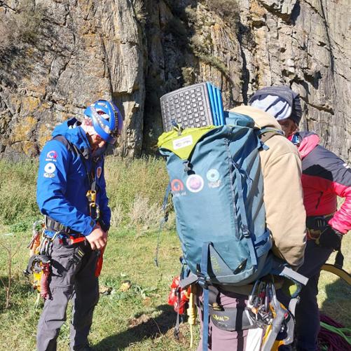 Trzy osoby przygotowują się do wspinaczki, mają na sobie uprzęże do wspinaczki, plecaki, a tle widać stromą ścianę
