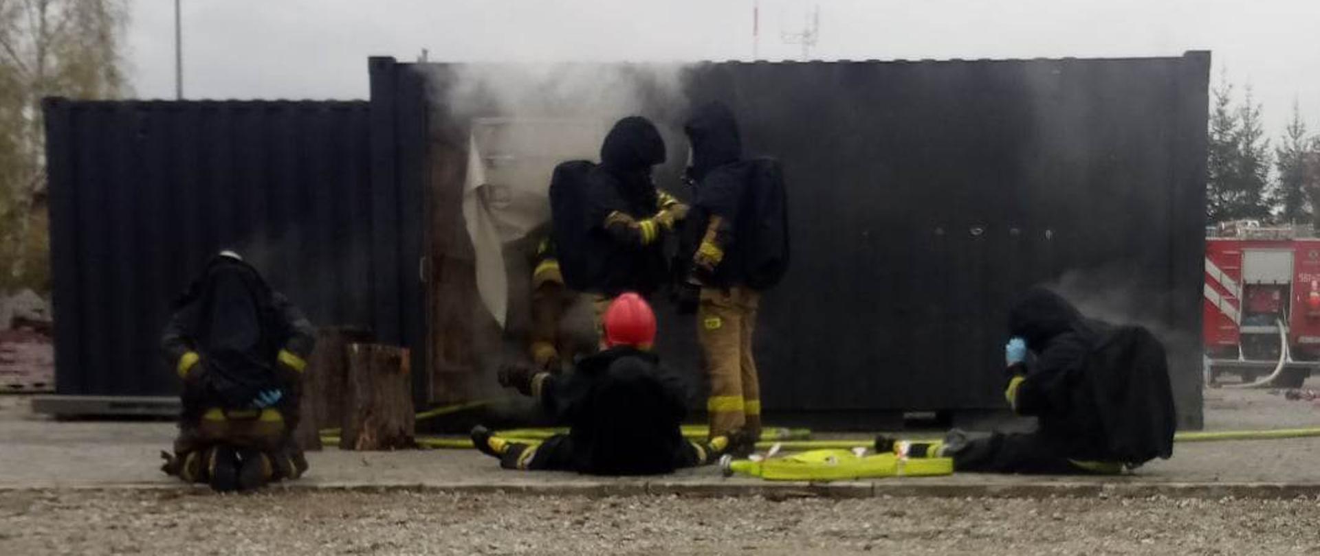 Na zdjęciu strażacy w ubraniach specjalnych oraz aparatach oddechowych po wyjściu z komory ogniowej. W tle komora oraz wydobywający się z niej dym