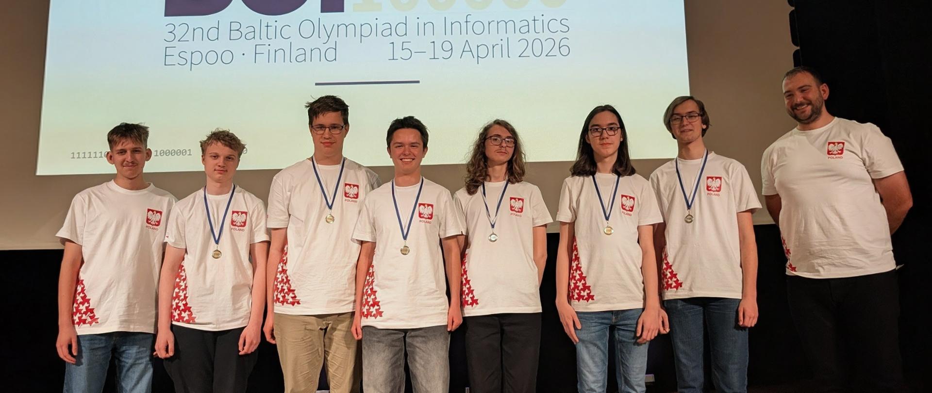 siedmiu młodych mężczyzn i jeden starszy stoi na tle dużego ekranu na którym wyswietalny jest napis BOI 32an Baltic Olympiad in Informatics Espoo Finland 15-19 April 2026