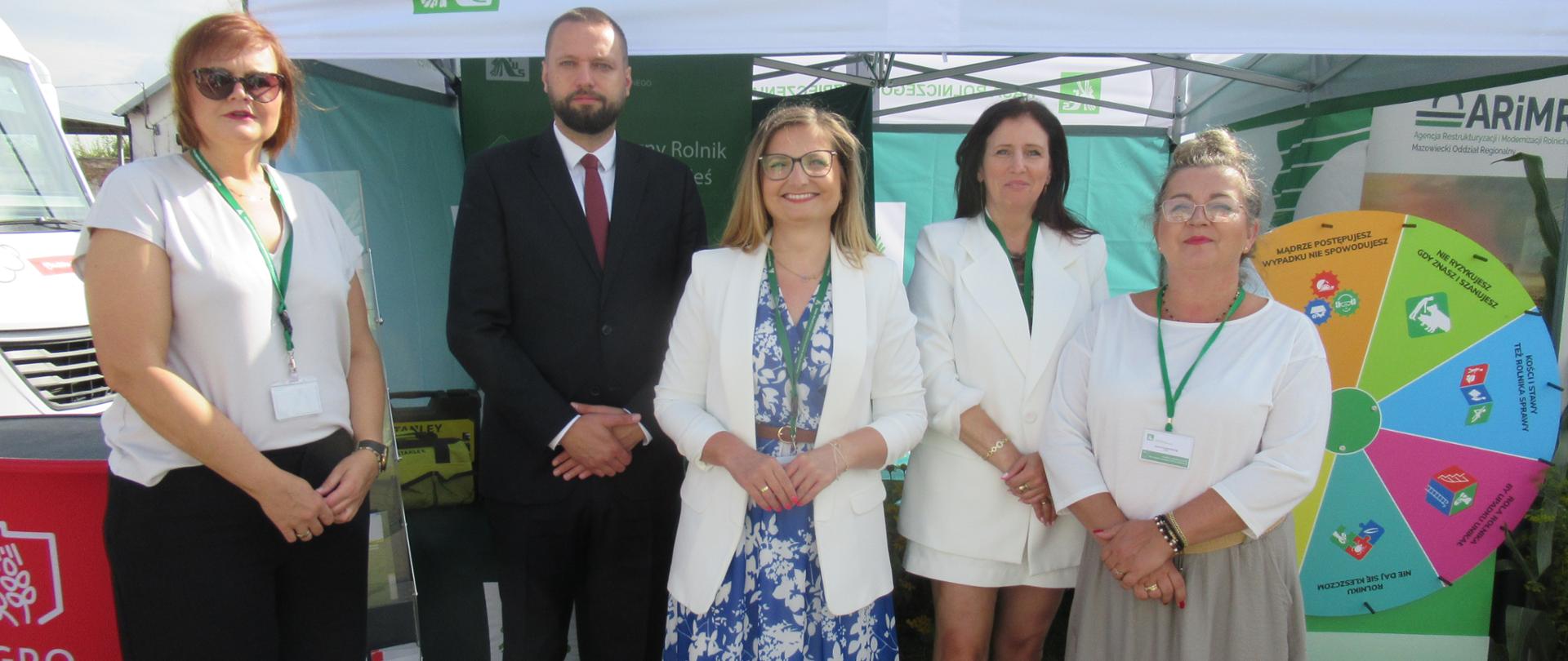 Cztery kobiety i mężczyzna pozują do zdjęcia przed stoiskiem pod namiotem z logo KRUS.