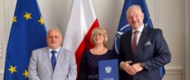 Mężczyzna w szarym garniturze, wicewojewoda oraz mężczyzna w ciemnym garniturze, wojewoda, pozują do zdjęcia z kobietą w granatowej sukience na tle flag: UE, Polski i NATO