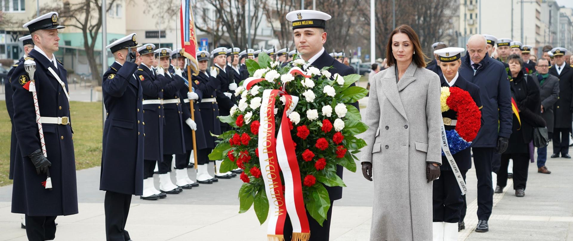 marynarz niesie wieniec biało czerwony, za nim idzie kobieta (wojewoda)